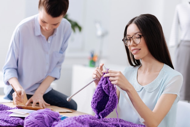 Affascinante giovane donna che lavora a maglia un cappello con fili viola, godendosi il processo mentre il suo collega crea modelli di vestiti