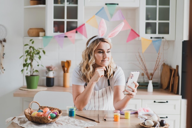 Affascinante giovane donna con il telefono in orecchie da coniglio dipingere le uova di Pasqua con vernici seduto al tavolo in cucina