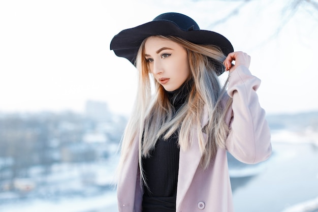 Affascinante giovane donna elegante in un cappello vintage in stile retrò in un cappotto rosa in un abito lavorato a maglia si trova in una calda giornata invernale