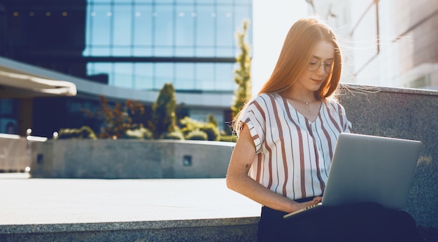 Affascinante giovane donna freelance che lavora al suo laptop all'aperto seduto sulle scale di un business center contro il tramonto.