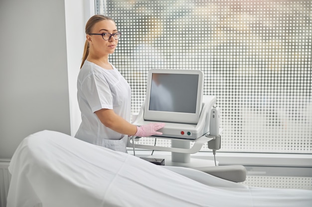 Affascinante giovane donna in guanti sterili in piedi da attrezzature moderne nel gabinetto di cosmetologia