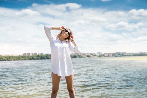 Affascinante giovane donna in un abito bianco godersi la natura in riva al lago in una giornata di sole. Servizio fotografico di moda alla moda