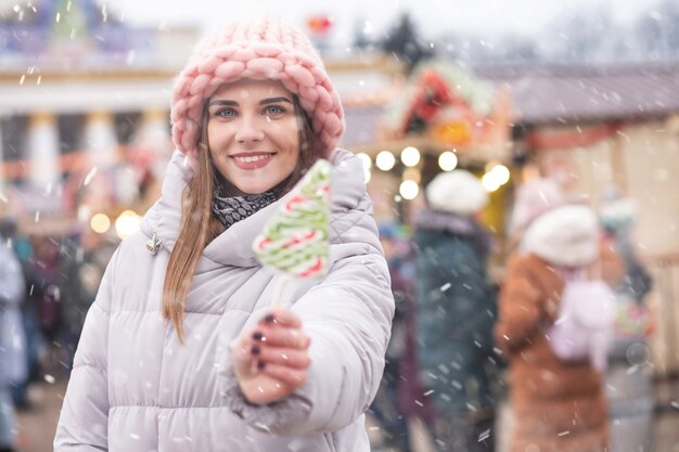 Affascinante giovane donna indossa un cappello rosa e un cappotto grigio che tiene caramelle sulla strada Fiera di Natale durante la nevicata. Spazio per il testo