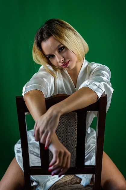 Affascinante giovane donna sexy sottile in abito bianco seduto in studio sulla sedia di legno su sfondo chiaro e ritratto in studio isolato sorridente
