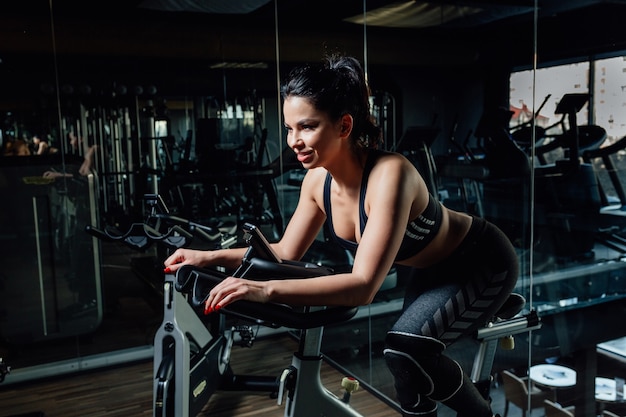 Affascinante giovane signora in abiti sportivi sorridente e seduto sulla moderna cyclette vicino allo specchio in elegante palestra