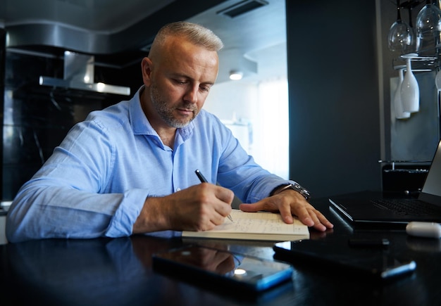 Affascinante imprenditore di mezza età, uomo d'affari che scrive su un taccuino, pianifica progetti e prende appunti in un diario, seduto davanti al monitor di un laptop a casa con interni eleganti.