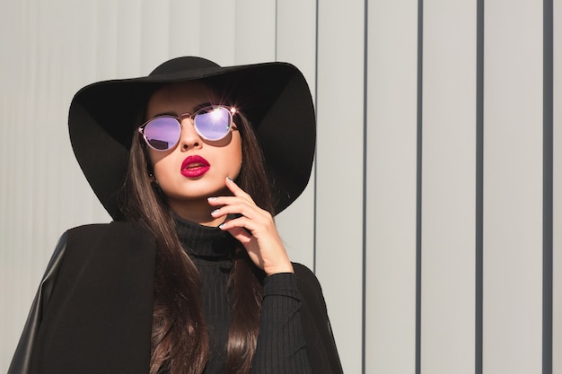 Affascinante modella bruna con occhiali a specchio e cappello a tesa larga in posa vicino alle persiane