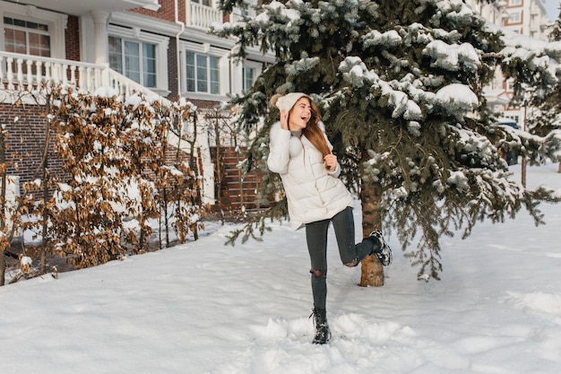 Affascinante ragazza alta in abiti invernali caldi che si divertono con la neve sullo sfondo dell'albero di abete nella mattina di sole. Modello femminile carino alla moda che esprime emozioni positive luminose in una giornata fredda..