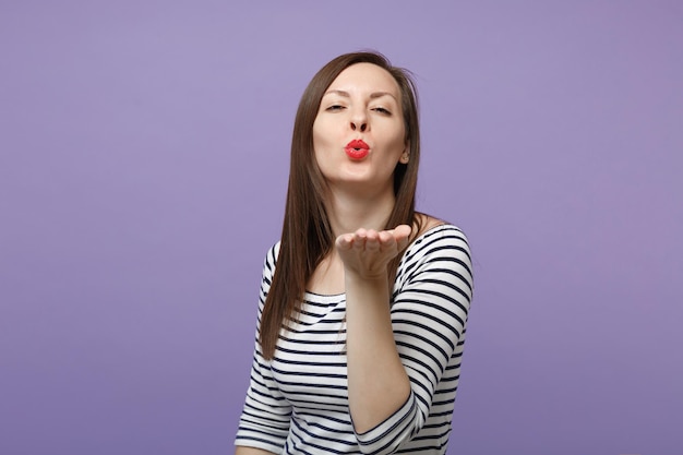 Affascinante ragazza carina giovane donna bruna in abiti casual a righe in posa isolata su sfondo viola viola ritratto in studio. Concetto di stile di vita delle persone. Mock up spazio di copia. Soffiando mandando un bacio d'aria.