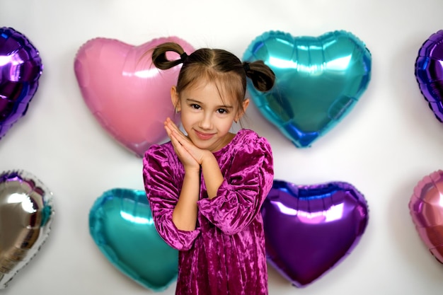 Affascinante ragazza con le code di cavallo in un abito viola di velluto si erge sullo sfondo di palloncini multicolori a forma di cuore per San Valentino, carina premendo le mani sulle guance