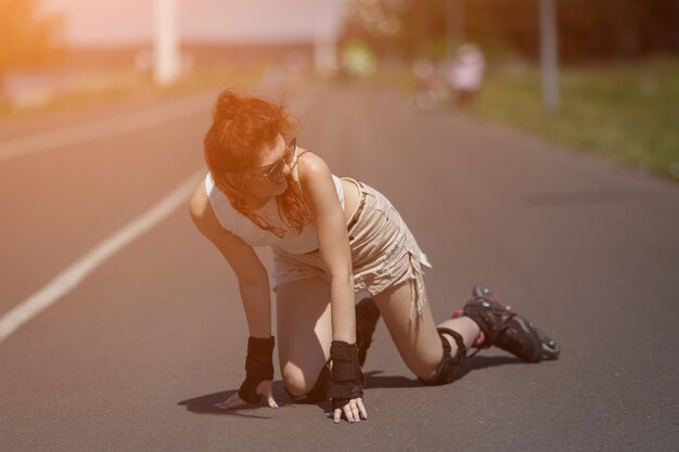 Affascinante ragazza dai capelli rossi in occhiali da sole e equipaggiamento protettivo cavalca su pattini a rotelle e cade per strada in una giornata di sole, luce solare