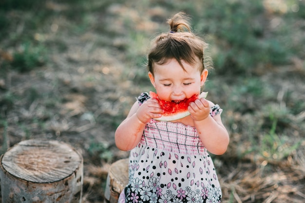 Affascinante ragazza di un anno e mezzo, mangia anguria all'aperto, nel villaggio contro gli alberi con una coda in testa