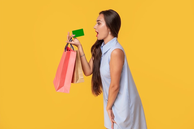 Affascinante ragazza sexy abbastanza attraente che mostra la carta di credito e tiene in mano molti pacchetti colorati, è comodo usare la carta di credito per lo shopping, in piedi su sfondo giallo