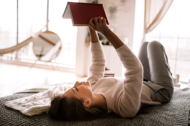 Affascinante ragazza vestita di maglione bianco e pantaloni legge un libro sdraiato sul letto con coperta grigia, cuscini bianchi e un regalo di Capodanno in una stanza di cory. .