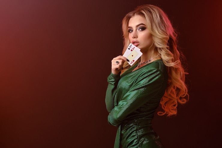 Foto affascinante signora bionda, trucco luminoso, in abito elegante verde e gioielli. piegò le mani, mostrando due assi, posando lateralmente su uno sfondo rosso da studio con retroilluminazione. gioco d'azzardo, poker, casinò. avvicinamento