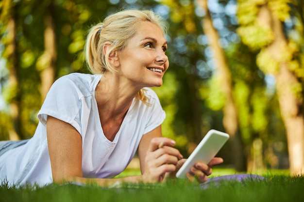 Affascinante signora che tiene il cellulare e sorride mentre riposa all'aperto sull'erba verde