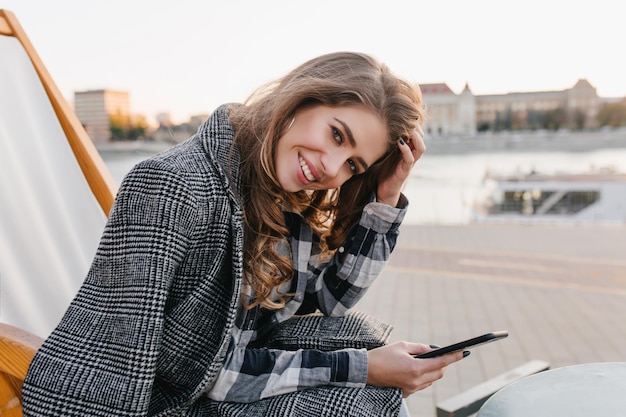 Affascinante signora dai capelli lunghi con il telefono in mano seduto su una poltrona reclinabile sullo sfondo della città. Modello femminile romantico in camicia a scacchi e cappotto grigio che riposa in chaise-longue in una giornata fredda.