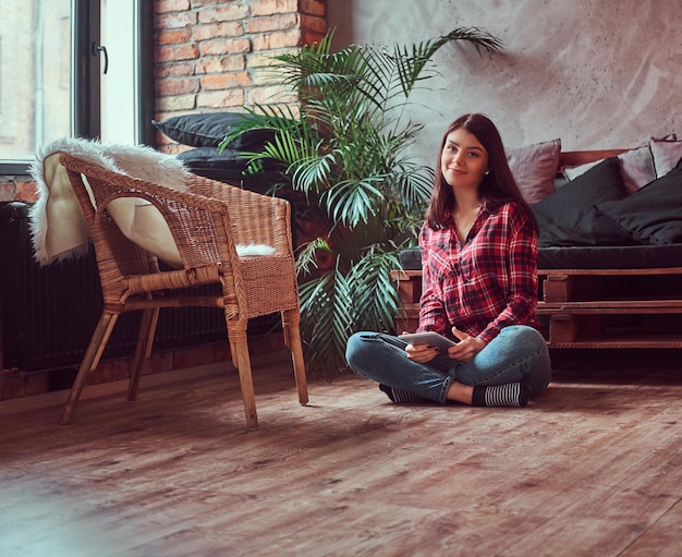 Affascinante studentessa bruna vestita con una camicia a quadri e jeans con in mano una tavoletta digitale seduta su un pavimento in una stanza con interni soppalcati, guardando la fotocamera.