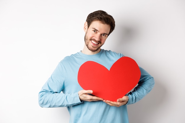 Affascinante uomo solitario che tiene il cuore rosso di San Valentino e sorridente, ti amo gesto, in piedi sul bianco