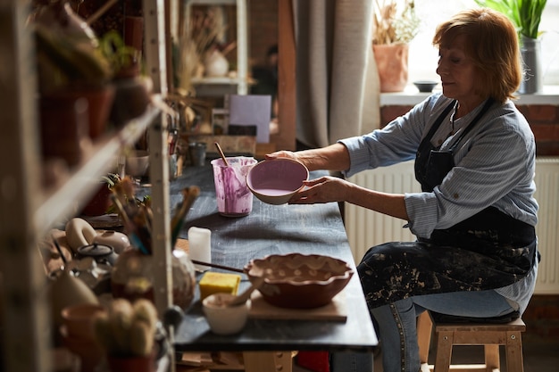 Affascinante vasaio seduto al tavolo e tenendo la ciotola di terracotta con vernice rosa mentre si lavora in studio di ceramica