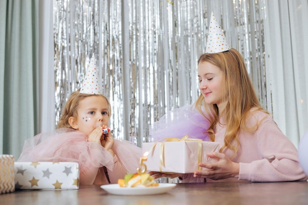 Affascinanti bambine dai capelli biondi con abiti da festa lilla e rosa con cappelli da festa, corno da festa e scatole regalo, sedute all'interno possibilmente in un caffè con sfondo decorato di lustrini argentati