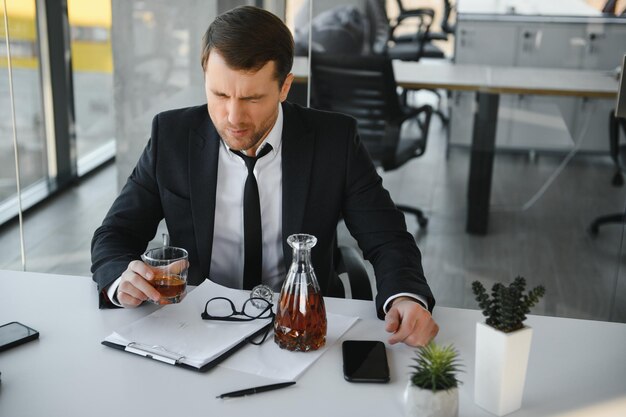 Affaticamento giovane uomo d'affari seduto sul posto di lavoro e tenendo la bottiglia di whisky bevendo alcol concetto di fallimento