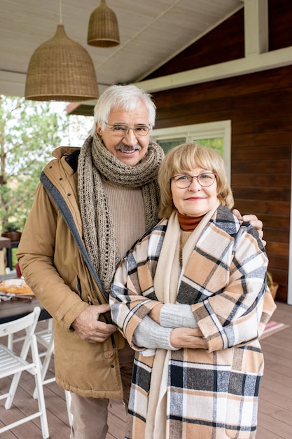 Affettuoso marito e moglie senior in un caldo abbigliamento casual che ti guardano con un sorriso mentre sono in piedi vicino alla loro casa contro il tavolo servito