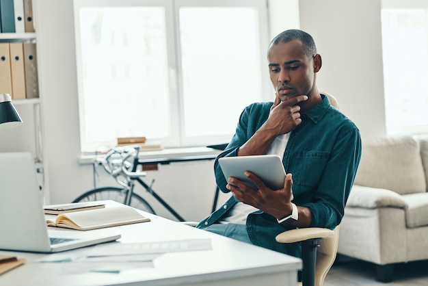 Affrettarsi a fare le cose. Riflessivo giovane africano in camicia che lavora utilizzando un tablet digitale mentre è seduto in ufficio