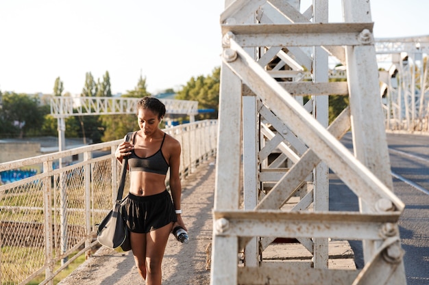 African American giovane donna in abbigliamento sportivo utilizzando auricolari e tenendo la bottiglia d'acqua mentre si cammina con la borsa sul vecchio ponte