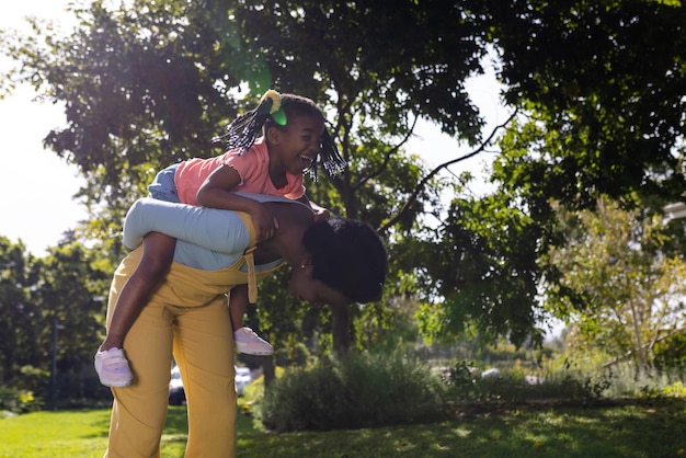 Afro-americana giocosa madre piggybacking figlia mentre si piega contro gli alberi nel parco. inalterato, stile di vita, famiglia, amore, solidarietà, infanzia, gioco, natura, estate e divertimento.