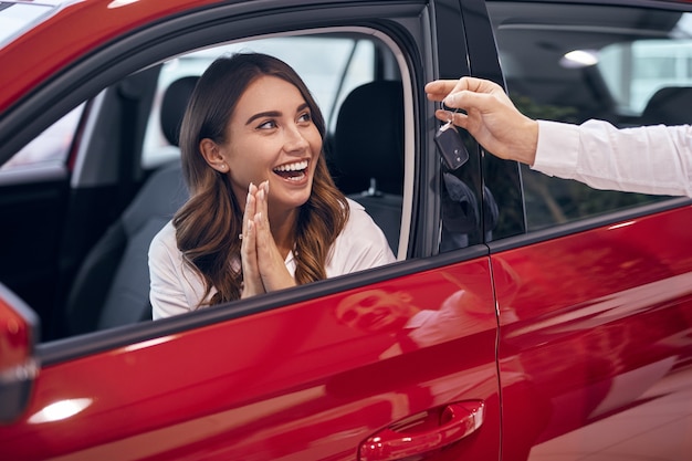 Agente dello showroom del raccolto che dà la chiave alla cliente femminile stupita che si siede in macchina e che applaude le mani