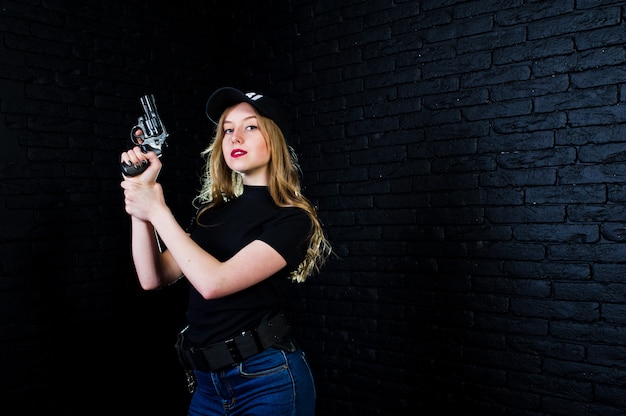 Agente femminile dell'FBI nel cappuccio e con la pistola contro il muro di mattoni scuro.