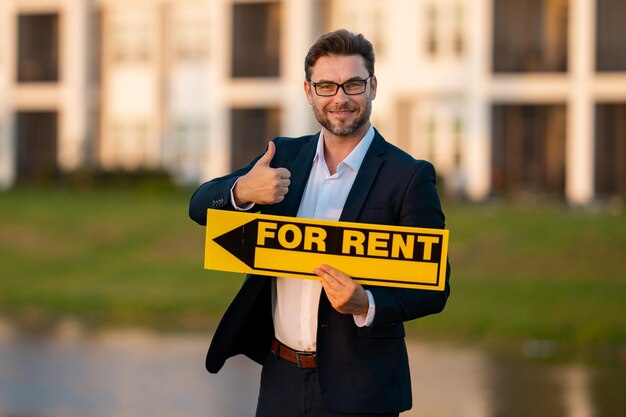 Agente immobiliare di successo in un abito in possesso di un cartello di affitto vicino a un nuovo agente immobiliare o immobiliare