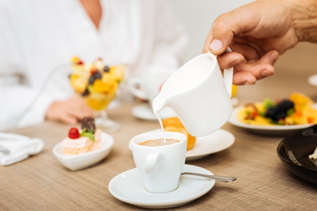 Aggiunta di creme. Uomo che mescola creme pesanti al suo caffè espresso caldo con una piacevole colazione