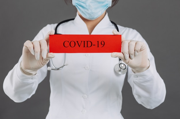 Aggiusti la donna con la maschera chirurgica che indica la carta rossa con il mesaage Coronavirus, isolato su fondo grigio