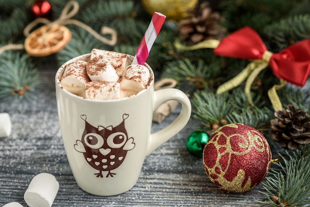 Aggredisca con cioccolata calda e la caramella gommosa e molle sul fondo di natale con i rami, i coni e le palle dell'abete sulla tavola di legno grigia.