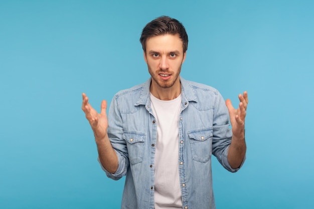 Aggressione e conflitto Ritratto di uomo irritato in camicia di jeans lavoratore alzando le mani e stringendo i denti con rabbia odio emozioni infastidite dal problema studio indoor girato isolato su sfondo blu