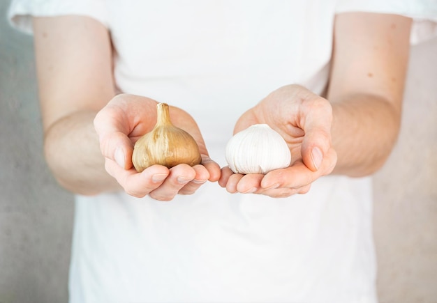 Aglio e aglio fermentato nelle mani dell'uomo Un uomo con una maglietta bianca su sfondo grigio tiene in mano aglio e bulbi di aglio nero nelle sue mani Cibo fermentato cura di sé alimentazione sana