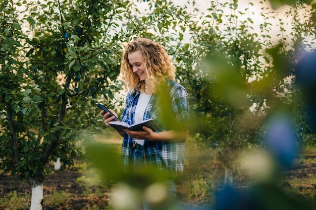 Agronomo donna che utilizza un telefono cellulare che prende appunti nel taccuino in piedi nel frutteto di prugne Gestione dell'agricoltura Concetto di tecnologia intelligente