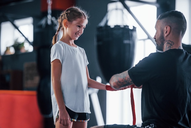 Aiutare a indossare le bende. Il giovane allenatore di boxe tatuato insegna a una bambina carina in palestra.