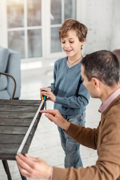 Aiutare papà. Bello piccolo ragazzo biondo attento che tiene un nastro di misurazione e misura il tavolo e suo padre che aiuta aiuta