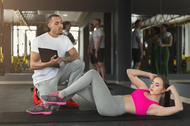 Aiuto allenatore di fitness con fare scricchiolii per atleta femminile al chiuso. Giovane uomo sportivo e donna che fanno sit-up sul tappeto. Stile di vita sano, ginnastica, concetto di perdita di peso