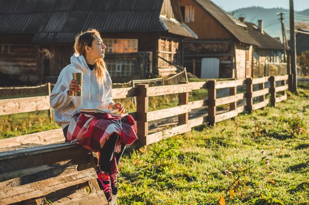 Al mattino la ragazza si siede su una panca di legno in mezzo alla natura, legge un libro e beve tè caldo da una tazza termica. Concetti Lettura in natura. Formazione all'aperto