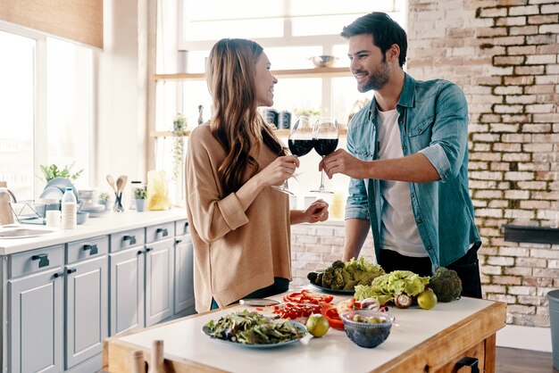 Al nostro amore! Bella giovane coppia che cucina la cena e beve vino stando in piedi in cucina a casa