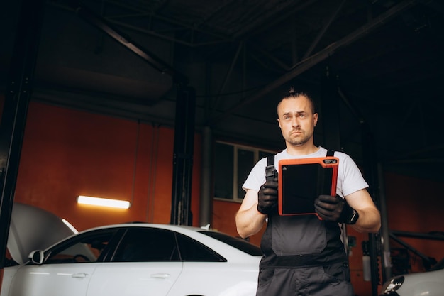 Al servizio auto un bel giovane meccanico automobilistico in uniforme sta usando un tablet digitale mentre il suo collega sta esaminando l'auto