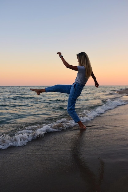 Al tramonto sulla spiaggia, una giovane donna si diverte e si tuffa vicino all'acqua in maglietta e jeans a piedi nudi