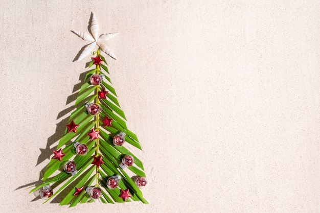 Albero di Natale fatto di brunch di palma verde con piccole decorazioni festive rosse sulla sabbia