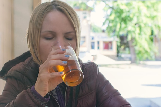 Alcolismo femminile Una donna in giacca beve birra da un bicchiere in un caffè di strada