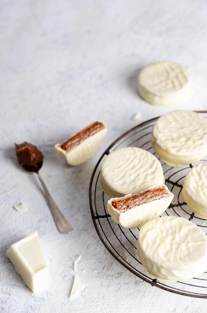 Alfajores al cioccolato bianco su una griglia scura con un pezzo di cioccolato bianco, su sfondo chiaro.