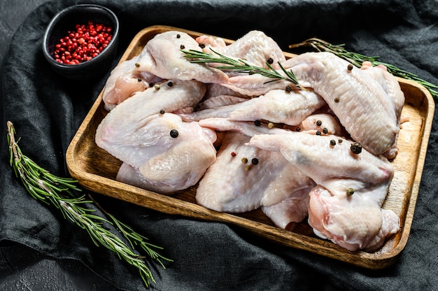 Ali di pollo crude in una ciotola di legno, cottura ingredienti rosmarino e pepe rosa. Fattoria biologica di carne. Vista dall'alto. Muro nero